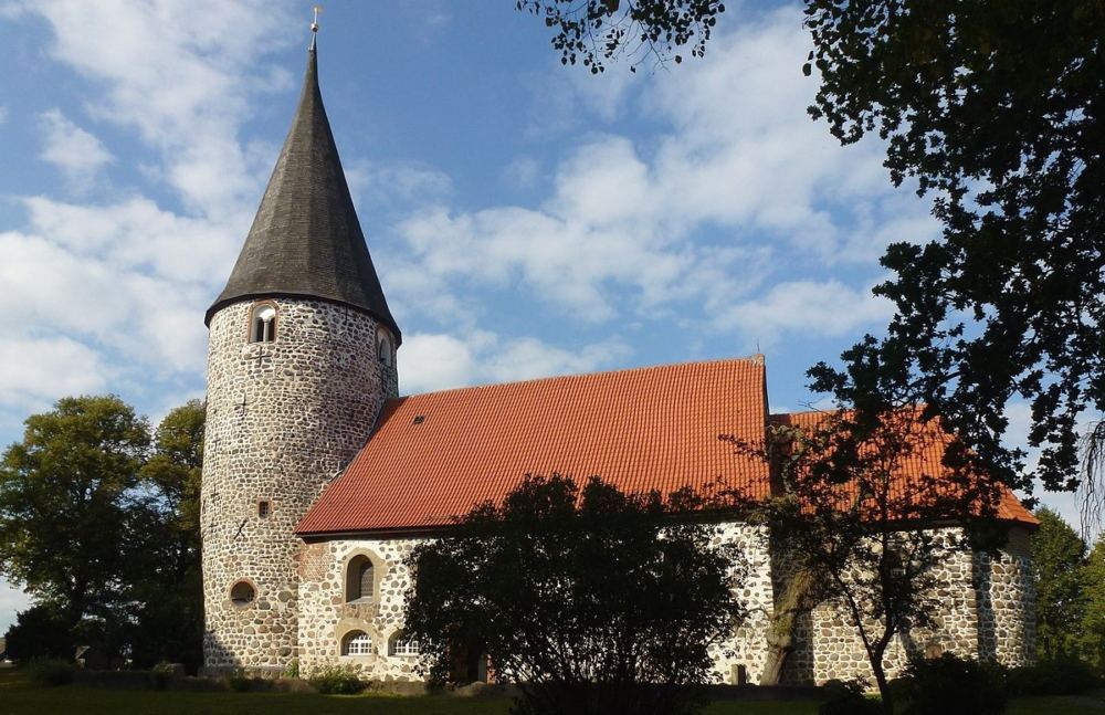 Vicelinkirche in Ratekau im Sonnenlicht, blauer Himmel mit Wolken, im Vordergrund schattige Büsche und Bäume