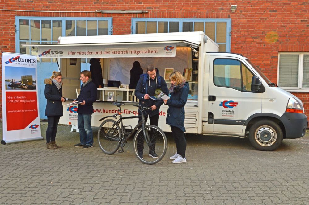 Infomobil zur Quartiersentwicklung. Weißer Lieferwagen als Informationsstand, davor reden Menschen miteinander.