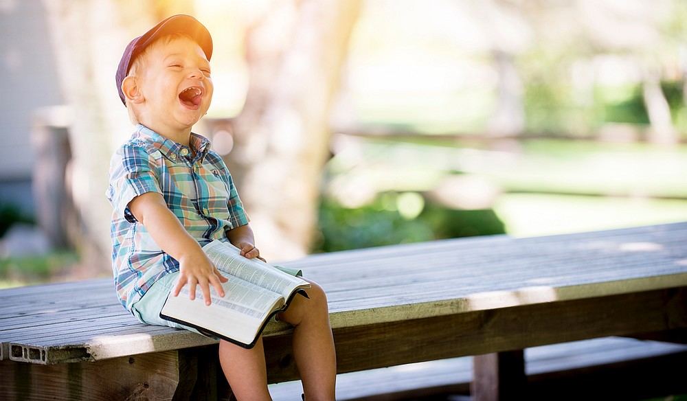 Ein Kind sitzt auf einer Bank, hält eine viel zu große Bibel und freut sich, ist stolz und lacht.