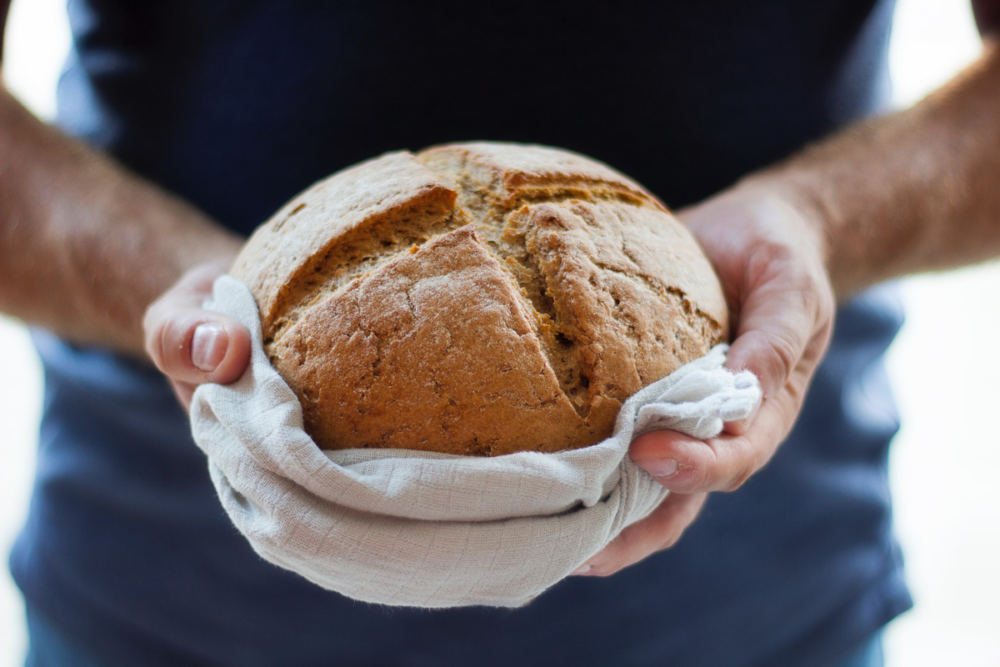 Hände halten ein frisches Brot. Die Krume ist kreuzförmig eingeschnitten.